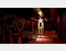 Dunkler, industrieller Innenraum mit Metalltischen, Wänden in Schwarz- und Rottönen, und neonroter Beleuchtung. An der Wand befindet sich eine große grafische Zeichnung, während in der Mitte ein grünes Bedienelement mit Hebeln und Schaltern steht.