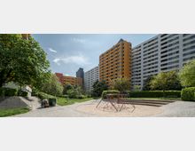 Ein Wohngebiet mit hohen, farbigen Apartmentgebäuden im Hintergrund, umgeben von Bäumen und Grünflächen. Im Vordergrund befindet sich ein kleiner Spielplatz mit einer Kletterpyramide aus Seilen auf einer Sandfläche.