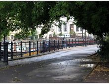 Ein Spazierweg entlang eines Kanals, gesäumt von Metallgeländern mit Bäumen im Vordergrund. Im Hintergrund sind Boote sowie historische Gebäude mit großen Fenstern sichtbar.