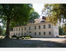 Ein zweistöckiges, cremefarbenes Herrenhaus mit symmetrisch angeordneten Fenstern und einem halbrunden Giebel über dem Eingangsbereich. Im Vordergrund befinden sich große Bäume und eine offene Rasenfläche mit vereinzelten Büschen.