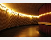 Ein geschwungener, moderner Innenraum mit senkrechten, strukturierten Wandpaneelen in warmem Gelbton. Der Boden besteht aus dunkel glänzenden Fliesen, und die rechte Wand ist mit Holzvertäfelung verkleidet.