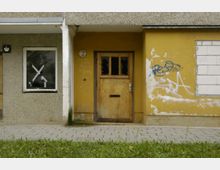 Fassade eines unsanierten Gebäudes mit einer gelben Wand, einem Holzeingang mit zerbrochenen Scheiben, Graffiti und Kritzeleien. Links befindet sich ein Fenster mit Klebeband und dahinter sichtbarem Innenraum; der Boden davor ist gepflastert, umgeben von einer Rasenfläche.