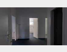 Ein Innenraum mit mehreren offenen weißen Türen, die zu unterschiedlichen Räumen führen. Der Boden ist mit dunkelblauem Teppich ausgelegt, an den Wänden befinden sich Heizkörper und ein teilweise sichtbares Fenster mit Jalousien.
