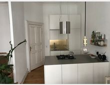 Moderne Küche mit weißen Schränken, einer Arbeitsplatte aus grauem Material und einer goldfarbenen Rückwand. Links ein Holzfußboden, rechts ein Wandregal mit Gläsern und Gewürzen, dazu eine hängende Glühbirne als Beleuchtung und eine Pflanze in der Ecke.