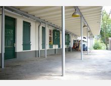 Überdachter Außenbereich eines Gebäudes mit weißen Wänden, grünen Türen und Fenstern, teilweise mit Gitterstäben. An der Wand hängen grüne Tafeln mit Speise- oder Warenangeboten, rechts stehen Menschen an einem Kiosk.