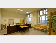 Ein schlichtes Büro oder Verwaltungszimmer mit Holzmöbeln, darunter ein Schreibtisch, eine Bank und ein Raumteiler. Die hohen Fenster mit Sprossenrahmen lassen Tageslicht herein, und an den Wänden befinden sich ein Heizkörper sowie Dekorationen wie Pflanzen und ein Kalender.