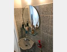 Kleines Badezimmer mit grauen Fliesen an der Wand, einem ovalen Spiegel darüber und einem kleinen Waschbecken mit geschwungenen Kanten. Über dem Waschbecken befindet sich ein Glastablar mit Kosmetikartikeln, darunter Parfüm und Cremes.