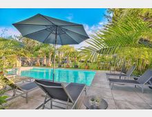 Ein Außenpool mit klarem, türkisfarbenem Wasser, umgeben von Liegestühlen, Sonnenschirmen und tropischen Palmen. Im Hintergrund befindet sich eine Rasenfläche mit weiteren Pflanzen und Sitzgelegenheiten.