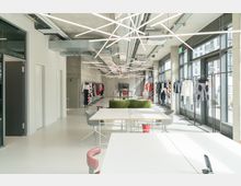 Offener, moderner Raum mit großen Fensterfronten, Kleiderständern mit farbiger Kleidung entlang der Wände und Schreibtischen im Vordergrund. Die Decke ist mit sichtbaren Lüftungsrohren und geometrischen, linearen Lichtinstallationen gestaltet.