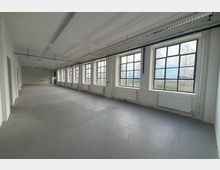 Großer, leerer Industrie- oder Lagerraum mit grauem Betonboden, weißen Wänden und sichtbaren Deckeninstallationen. Entlang einer Seite verläuft eine Reihe großer, schwarz gerahmter Sprossenfenster mit Heizkörpern darunter und Blick ins Freie.