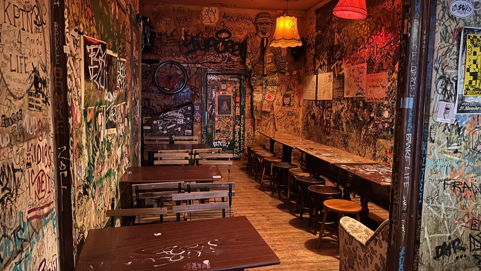 Schmaler Innenraum einer Bar oder eines Cafés mit Holzdielen, langen Holztischen und Hockern sowie mehreren Hängelampen mit farbigen Stoffschirmen. Wände und Türflächen sind dicht mit Graffiti, Stickern und Plakaten bedeckt, wodurch ein stark urbaner, dekorierter Look entsteht.