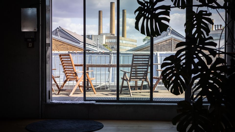 Blick aus einem Innenraum mit Topfpflanze auf eine Terrasse mit Holzmöbeln, Glasgeländer und Industriegebäuden mit Schornsteinen im Hintergrund.
