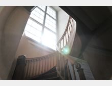 Holztreppe mit geschwungenem Geländer in einem historischen Gebäude, aufgenommen von unten mit Blick auf ein großes, von Sonnenlicht durchflutetes Fenster.