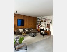 Wohnzimmer mit modernem Design, bestehend aus einer braunen Eckcouch, einem schwarz-weißen Sitzpouf und einem schwarzen Sessel. An einer Wand aus Holzpaneelen hängt ein dunkelblaues Bild, daneben befindet sich ein Regal mit Büchern und Dekorationsgegenständen. Ein heller Teppich bedeckt den Betonboden, und im Vordergrund steht eine grüne Zimmerpflanze.