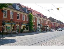 Straßenszene mit einer Reihe von Gebäuden aus rotem Backstein mit Satteldächern, einigen Fenstern und Ladengeschäften im Erdgeschoss, darunter ein Café mit Außensitzplätzen und einem Modegeschäft. Im Vordergrund sind eine gepflasterte Straße, Straßenbahnschienen und einige Fußgänger sowie Fahrräder zu sehen.