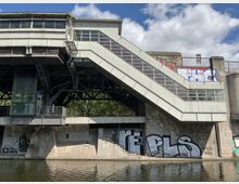 Ansicht eines erhöhten Bahnhofs oder einer Brücke mit Treppe und Aufzug, die über einen Fluss führt. Die Betonwände darunter sind mit Graffiti bedeckt, und das Bauwerk besteht aus Metall- und Glasstrukturen.