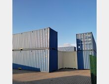 Gestapelte Frachtcontainer in Blau und Weiß stehen auf einer gepflasterten Fläche unter klarem, blauem Himmel. Im Vordergrund ist ein Maschendrahtzaun sichtbar.
