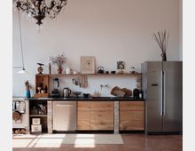 Rustikale Küche mit Holzmöbeln, schwarzer Arbeitsplatte und freigelegten Ziegelsteinen. An der Wand hängen Messer und Regale mit Geschirr, ein großer silberner Kühlschrank steht rechts, während links ein alter Küchenofen sichtbar ist; eine Kristalllampe hängt von der Decke.