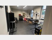 Ein heller Tätowierladen mit mehreren Liegen, schwarzen Möbeln und Wanddekorationen wie Bilderrahmen und Kunstwerken. Große Fenster lassen Tageslicht herein, und im Raum stehen Arbeitsleuchten und Werkzeuge.