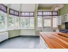 Heller Küchenraum mit großen, umlaufenden Fenstern und halbtransparenten Raffrollos, hellgrünen Wänden und grauem Fliesenboden. Rechts sind weiße Küchenschränke, ein Herd sowie eine breite Holzarbeitsplatte zu sehen, daneben eine Glastür nach außen.