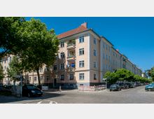 Eckhaus mit heller Fassade und roten Ziegeldächern in einem Wohngebiet, umgeben von gepflasterten Straßen und Bäumen. Im Vordergrund sind parkende Autos, Gehwege und ein Straßenschild zu sehen.