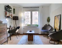 Helles Wohnzimmer mit großen Fenstern und weißem Vorhang, das Tageslicht hereinlässt. Der Raum ist mit einem Teppich, einer Holzkommode, einem niedrigen Tisch und modernen Sitzmöbeln eingerichtet, ergänzt durch Pflanzen und Wanddekoration.