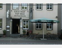 Fassade eines Gasthofs mit der Aufschrift „Gasthof Stadt Berlin, Inh.: Franz Heinrich“. Vor dem Eingang stehen Tische und Stühle unter einem grünen Sonnenschirm mit der Aufschrift „Diebels“, daneben ein Kind sowie Pflanzen in Töpfen.