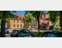 Straßenecke in einer kleinen Stadt mit gepflasterter Fahrbahn, geparkten Autos und großen Straßenbäumen im Vordergrund. Im Hintergrund stehen zweigeschossige Altbauten mit roten Ziegeldächern und hellen Fassaden, darunter ein Gebäude mit der Aufschrift „Ristorante Pizzeria“, sowie Ampeln und Verkehrszeichen an der Kreuzung.