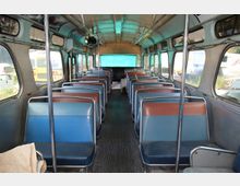 Innenraum eines alten Linienbusses mit zwei Reihen aus gepolsterten Sitzen in Blau und Braun. Die Wände und Decke sind in Metalloptik gehalten, große seitliche Fenster lassen Licht herein.