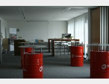 Ein heller Veranstaltungsraum mit grauem Teppichboden, mehreren roten Metallfässern mit "Total"-Logo, die als Stehtische genutzt werden, und einem langen Holztisch mit weißen Barhockern. Im Hintergrund stehen Tische mit Getränken, Gläsern und Snacks vor großen Fenstern mit Jalousien.