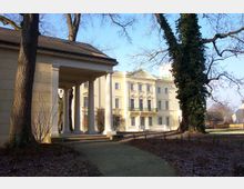 Gelber, klassizistischer Prachtbau mit Säulengang im Vordergrund, umgeben von Bäumen und einem parkähnlichen Gelände. Im Hintergrund sind das Hauptgebäude mit symmetrischer Fassade und ornamentierten Giebeln sowie ein klarer Himmel zu sehen.