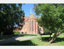 Backsteingebäude einer historischen Kirche mit hohen Spitzbogenfenstern und einem grünen Turm vor einem blauen Himmel, umgeben von Rasenflächen und Bäumen; im Vordergrund steht eine Holzbank unter einem kleinen Baum.