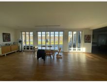 Heller Wohnraum mit Holzboden, einem Esstisch und Stühlen in der Mitte. Große Fensterfront mit Blick auf eine Terrasse, einen Garten und einen See im Hintergrund.