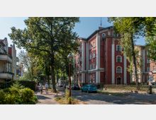 Straßenszene mit einem historischen Backsteingebäude rechts, verziert mit weißen Säulenelementen und Rundbogenfenstern. Links befinden sich Wohnhäuser mit Balkonen, die Straße ist gesäumt von Bäumen und geparkten Autos, und im Vordergrund spazieren zwei Personen.