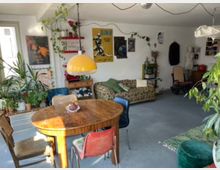 Eclectic living room with a round wooden dining table and mixed chairs in the foreground, a yellow dome pendant lamp, and a patterned sofa against a white wall covered with posters. The space features many potted and hanging plants, open shelving, and a carpeted floor with assorted vintage furniture and decor.