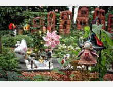 Gräberfeld in einem Garten mit bunten Dekorationen, darunter Blumen, Figuren und ein Stoffpüppchen, das an einer Laterne hängt. Im Hintergrund stehen rotbraune, gebogene Steinskulpturen und Bäume.