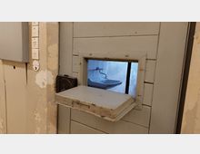 Metalltür mit einer kleinen rechteckigen Durchreiche, die nach unten geöffnet werden kann; durch die Durchreiche ist ein Waschbecken in einem gekachelten Raum sichtbar. Die Wand neben der Tür ist abgenutzt und mit Schaltern versehen.