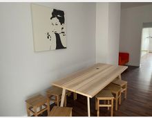 Minimalistisch eingerichteter Raum mit einem hellen Holztisch, umgeben von passenden Holzhockern, auf einem dunklen Holzboden. An der weißen Wand hängt ein schwarz-weißes Porträtbild, im Hintergrund sind ein orangenes Sofa und ein anderer Raum sichtbar.