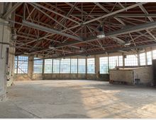 Industriehalle mit leerem, betoniertem Boden, großen bodentiefen Fenstern und einer Dachkonstruktion aus Stahlträgern und Holz. Die Halle wirkt ungenutzt und weist an den Wänden sichtbare Abnutzungsspuren auf.