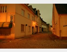Eine Kopfsteinpflasterstraße in einer kleinen Stadt, flankiert von mehrstöckigen Wohn- und Geschäftsgebäuden mit schlichten Fassaden. Straßenlaternen werfen ein warmes orangefarbenes Licht, während der Himmel trüb wirkt.