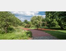 Ein Landschaftspark mit einem gepflasterten Weg, der durch eine grüne Wiese und üppige Vegetation führt. Rechts steht eine Holzschaukel, umgeben von Rasen und Bäumen, unter einem bewölkten Himmel.
