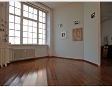 Ein heller Raum mit Holzboden und hohen Decken, geprägt durch ein großes Fenster mit Sprossen, das viel Tageslicht hereinlässt. An den weißen Wänden hängen mehrere gerahmte Kunstwerke, und unter dem Fenster befindet sich ein Heizkörper.