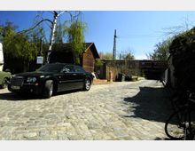 Kopfsteinpflasterstraße mit einem schwarzen Auto geparkt im Vordergrund. Im Hintergrund befinden sich Holzhütten, eine Eisenbahnbrücke und ein Strommast, umgeben von Bäumen und Vegetation.