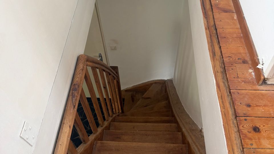 Blick von oben in ein schmales Treppenhaus mit steiler, hölzerner Treppe und Geländer aus Holz. Weiße Wände und ein Holzfußbodenrahmen um die Treppenöffnung prägen den Innenraum.