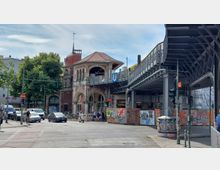 Straßenszene mit einem historischen U-Bahnhof aus Backstein und Arkaden rechts im Bild, über dem eine erhöhte U-Bahnlinie verläuft. Im Vordergrund sind eine Kreuzung, Fahrzeuge, Fußgänger sowie Baustellenzäune mit Graffiti zu sehen.