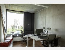 Modernes Büro mit minimalistischer Einrichtung, darunter zwei Schreibtische mit Computern und Schreibtischlampen, ein großes Fenster mit Blick auf Gebäude und Bäume sowie Betonwände und eine Holzfußbodenfläche. Ein Pinnwand und geöffnete Unterlagen befinden sich ebenfalls im Raum.