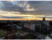 Städtische Skyline bei Sonnenuntergang mit dicht gedrängten Gebäuden und Wohnhäusern im Vordergrund, einer parkähnlichen Grünfläche mit einer historischen Fassade sowie einem wolkenbedeckten Himmel im Hintergrund.