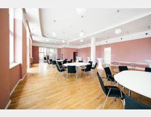 Moderner Veranstaltungsraum mit runden und rechteckigen Tischen, umgeben von schwarzen Stühlen. Der Raum hat eine helle Holzoptik, rosa gestrichene Wände, große Fenster und weiße Deckenlampen in kugelförmigem Design.