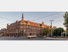 Backsteingebäude im neugotischen Stil mit vielen Spitzbögen und einem kleinen Turm; im Vordergrund eine Straßenbahnhaltestelle, eine Straßenbahn und vereinzelte Fußgänger, rechts ein kleiner Park mit Bäumen.