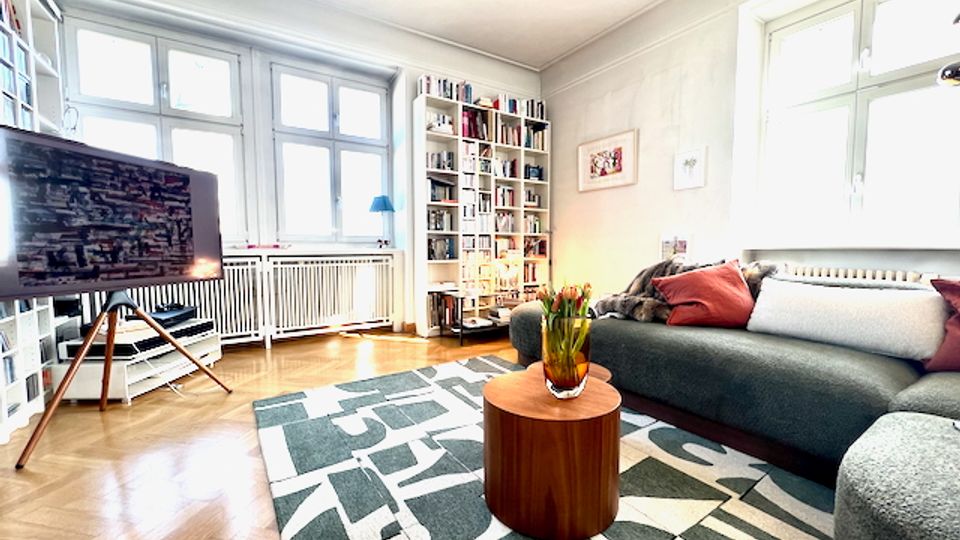 Helles Wohnzimmer mit großen Sprossenfenstern, Parkettboden und gemustertem Teppich, in der Mitte ein runder Couchtisch vor einem grauen Sofa. An den Wänden stehen hohe Bücherregale, links befindet sich ein Fernseher auf einem Stativ neben einem Heizkörper.